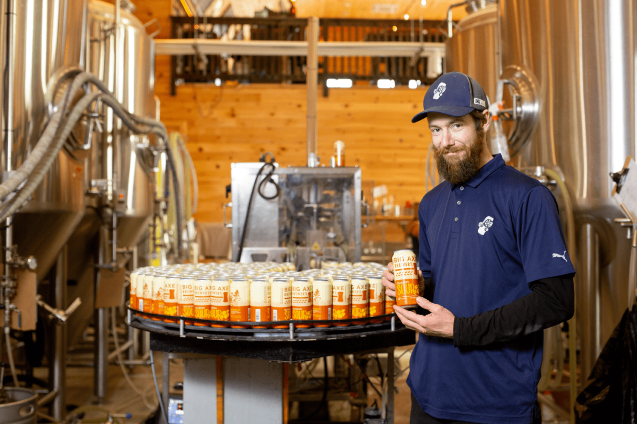 Craft Coast Packaging team member standing in front of several Big Axe Brewery printed cans. The team member is wearing a navy tshirt over a black long-sleeved shirt and is wearing a navy hat with the Craft Coast Packaging logo on it.